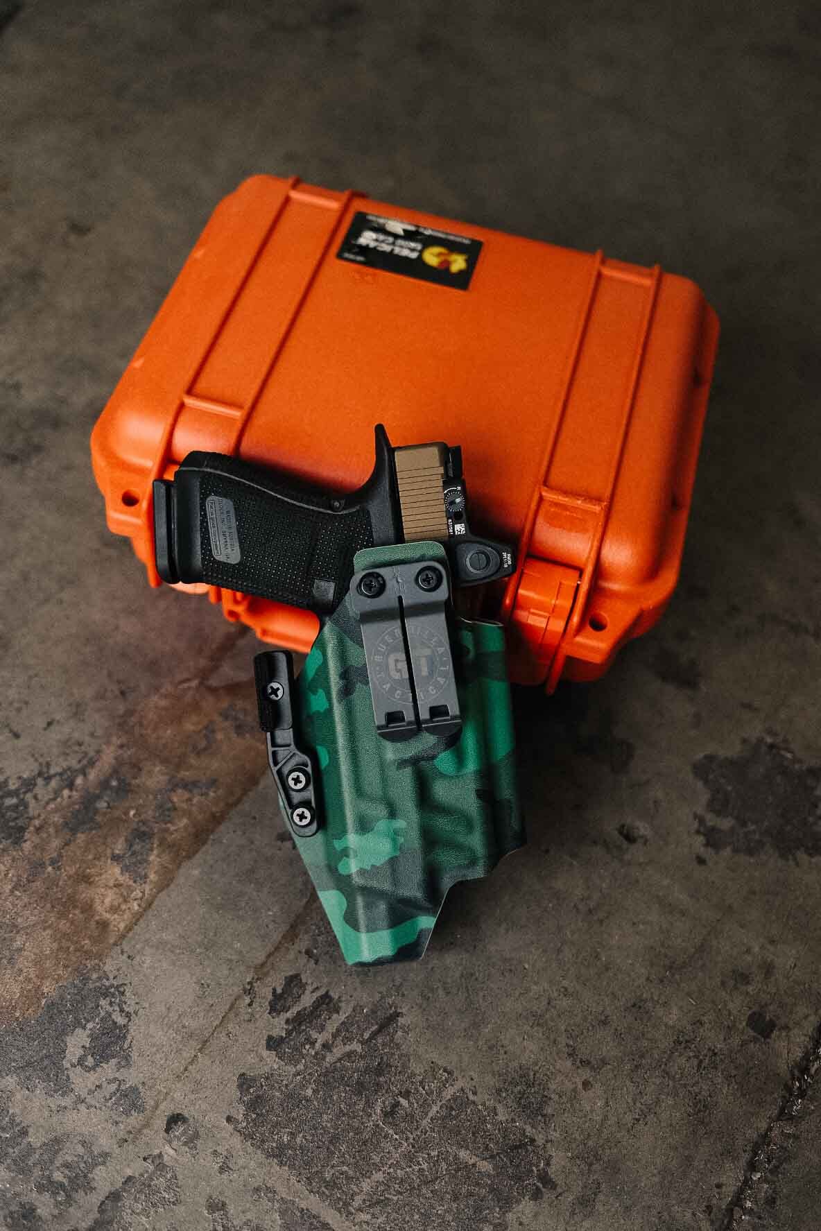 A handgun with a tan slide and black grip securely holstered inside a green camouflage Guerrilla Tactical Kydex IWB holster featuring a dual-clip attachment with the circular GT logo, resting on an orange Pelican hard case on a concrete shop floor
